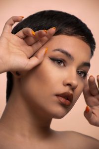 A striking close-up portrait of a woman with bold makeup and orange nails against a neutral background.