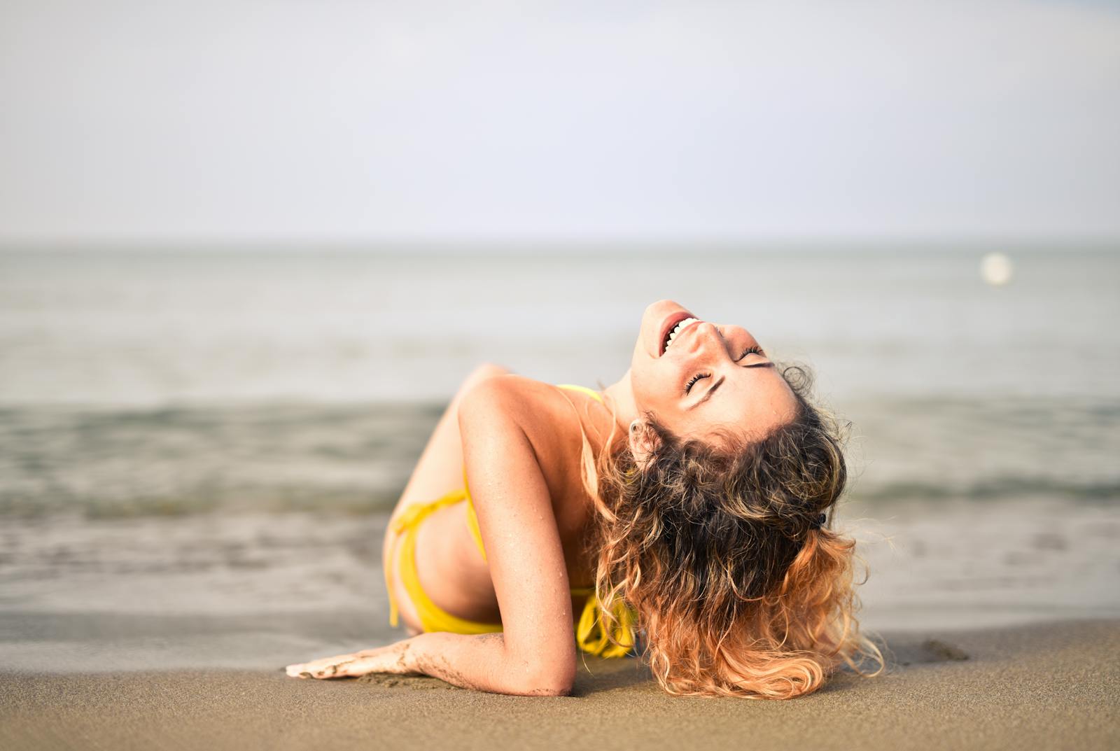 Woman relaxing and laughing on a sandy beach by the ocean, enjoying a sunny summer day in a swimsuit.