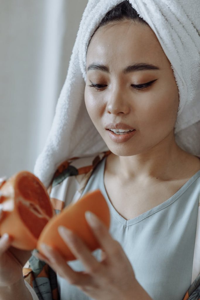 Young woman with a towel on her head enjoys a fresh grapefruit indoors, embodying relaxation and self-care.