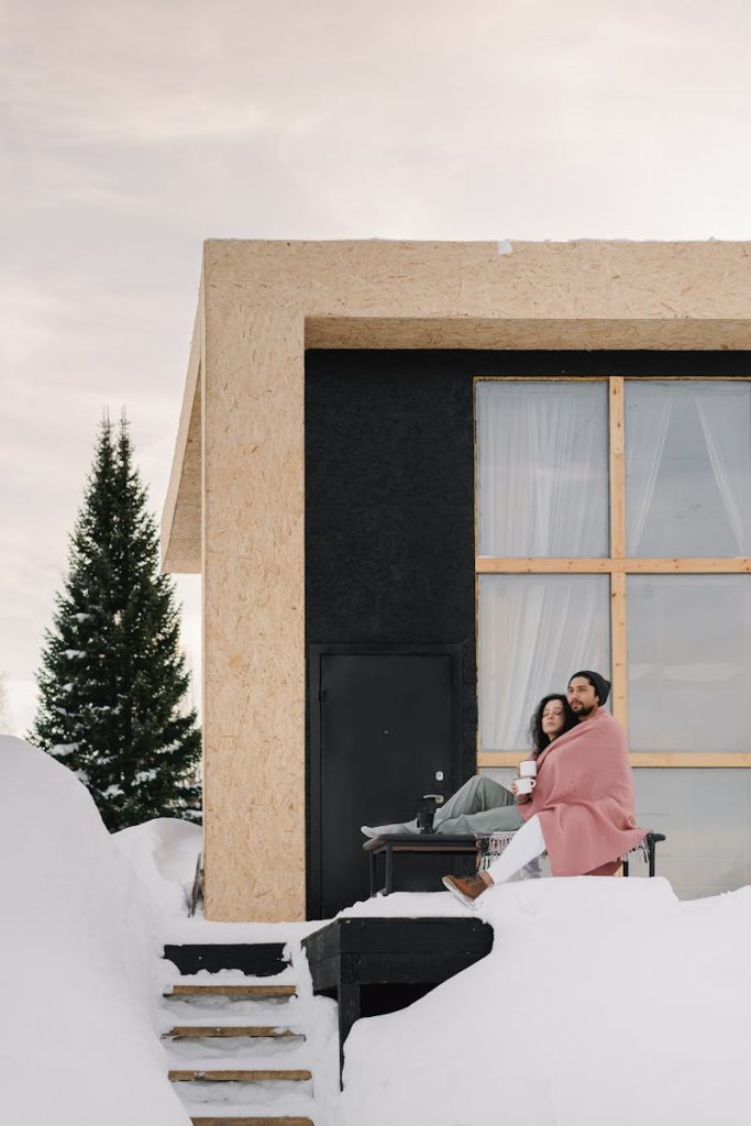 Couple wrapped in blanket enjoying winter outside a modern cabin on snowy day.
