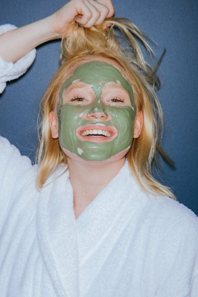 Woman in a bathrobe smiling while applying a green mud mask for skincare treatment.