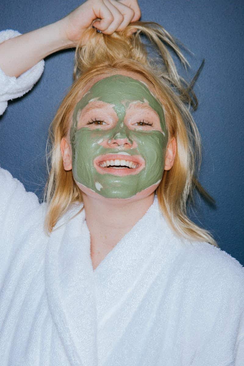 Woman in a bathrobe smiling while applying a green mud mask for skincare treatment.